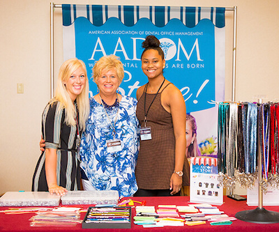 Three women standing in front of a AADOM sign and behind a desk
